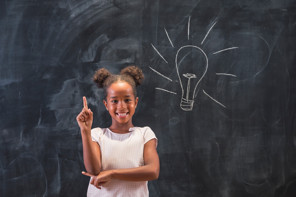 Elementary school girl standing in front of a chalkboard in classroom, happy after coming up with an idea, holding index finger up with light bulb drawn on the blackboard behind her